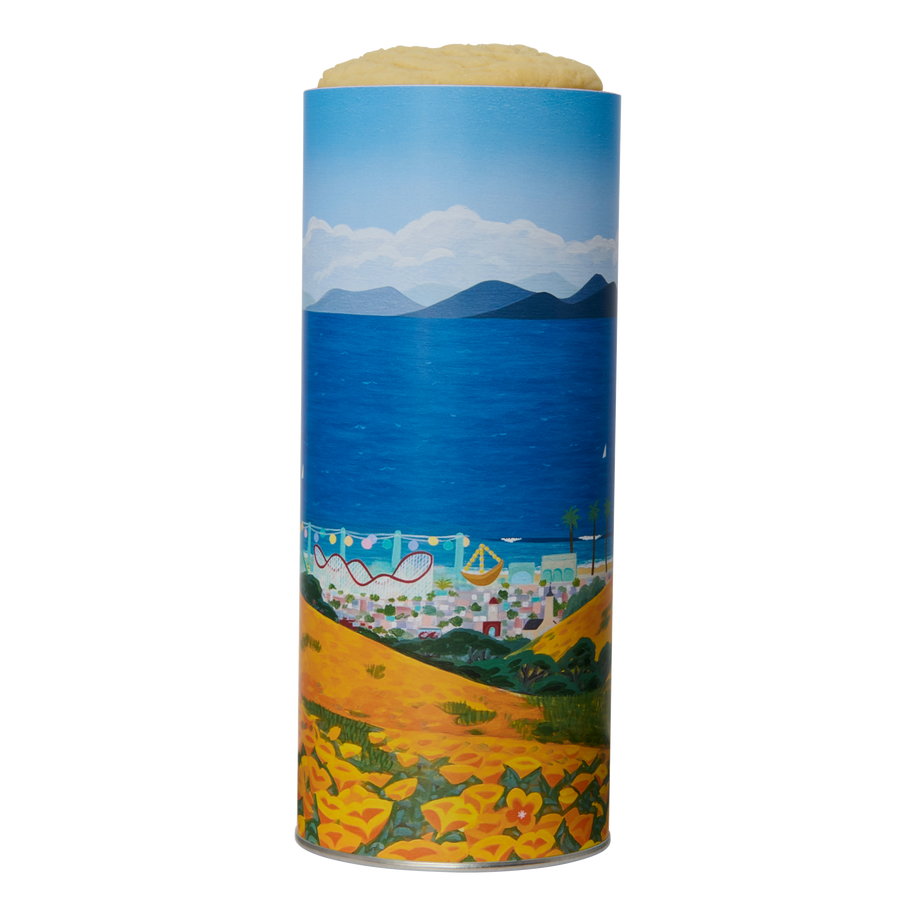 A cylindrical tin of cookies with a colorful coastal landscape illustration, featuring a blue sea, mountains, beach, and yellow flowers.